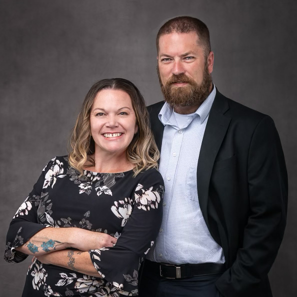 Chris and Shanna Caldwell, smiling and posing together for their profile picture. Chris, wearing a smart, dark-colored suit and tie, stands on the left, while Shanna, elegantly dressed in a light-colored blouse, stands on the right. They appear professional and approachable, embodying the leadership behind their company.